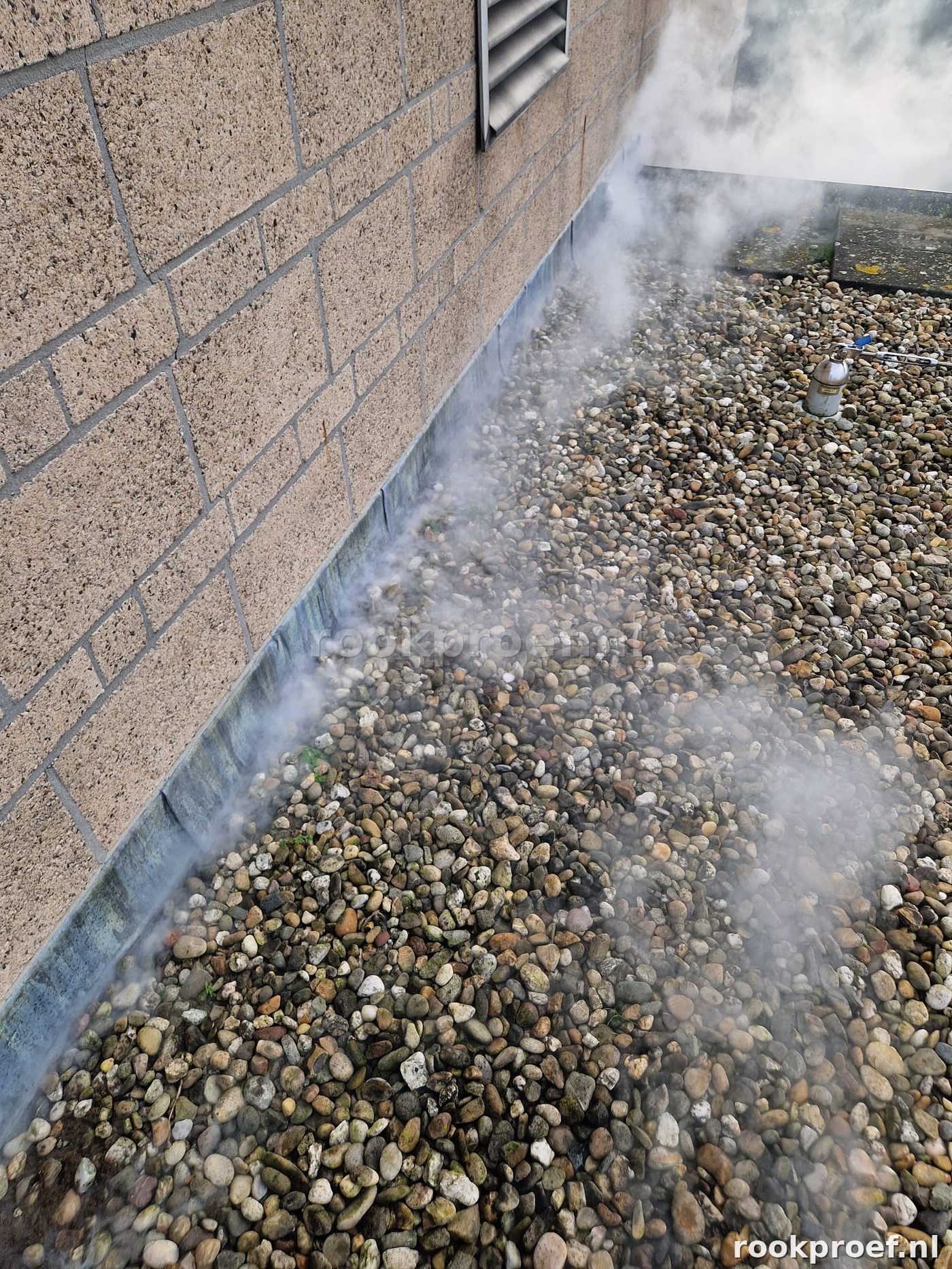 Rookgas ontsnapt zichtbaar langs dakrandaansluiting bij gevel op grindbed — lekpunt gevonden