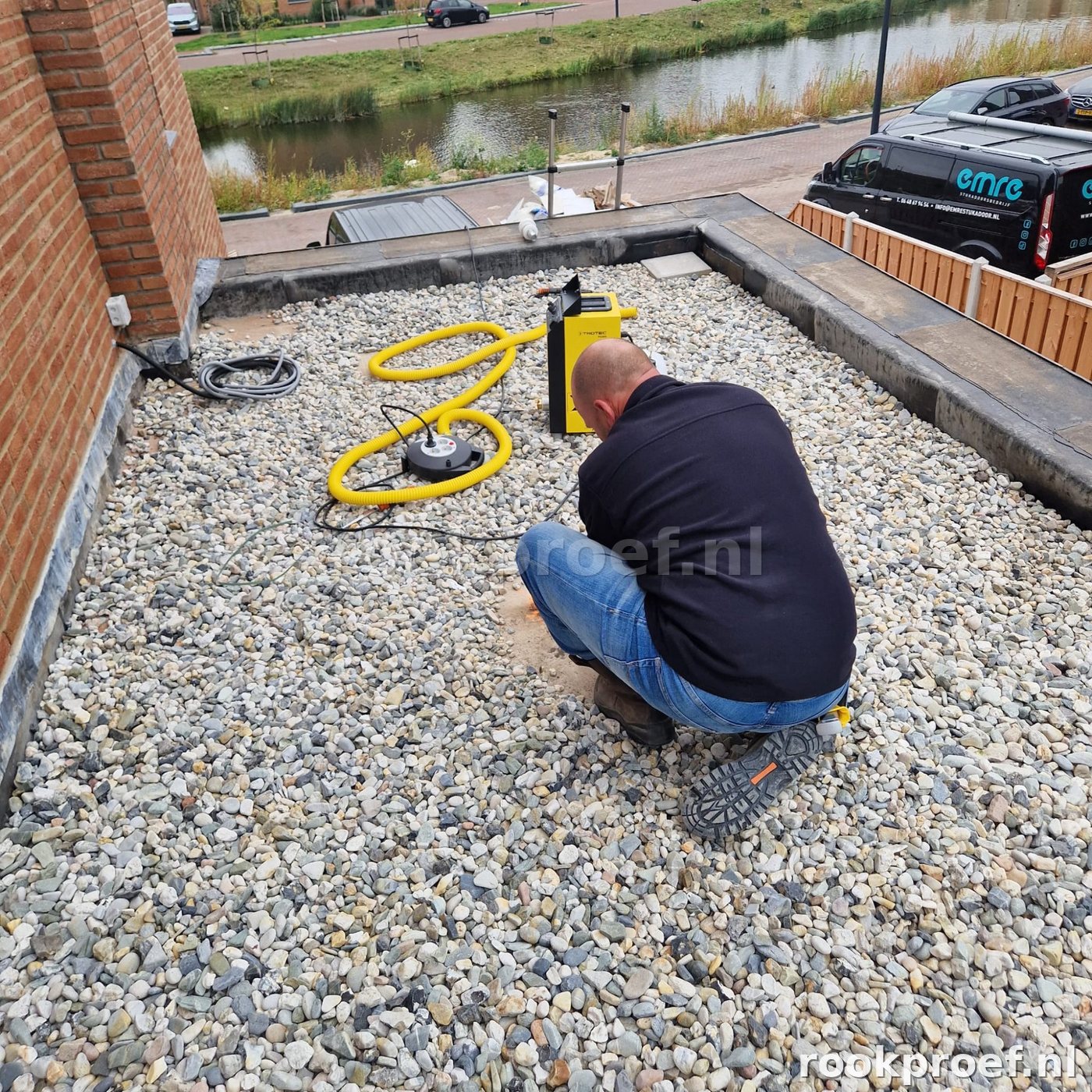 Rookproef Nederland technicus knielt op grindlaag — Trotec rookmachine met gele slang daklekkage test
